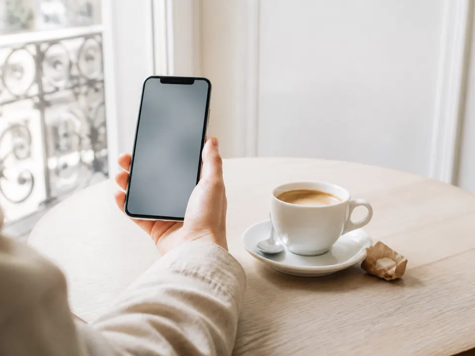 Main tenant un smartphone à l'écran flou, sur une table éclairée par la fenêtre