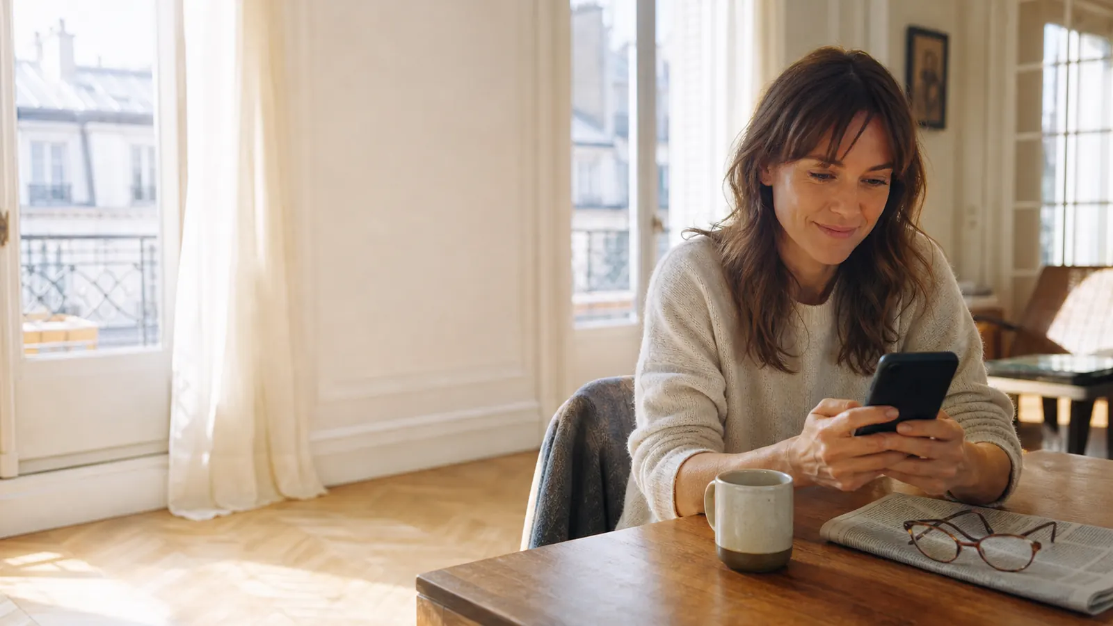 Vue large d'un salon lumineux aux hautes fenêtres : une femme assise à une table en bois lit un message sur son téléphone, un sourire retenu